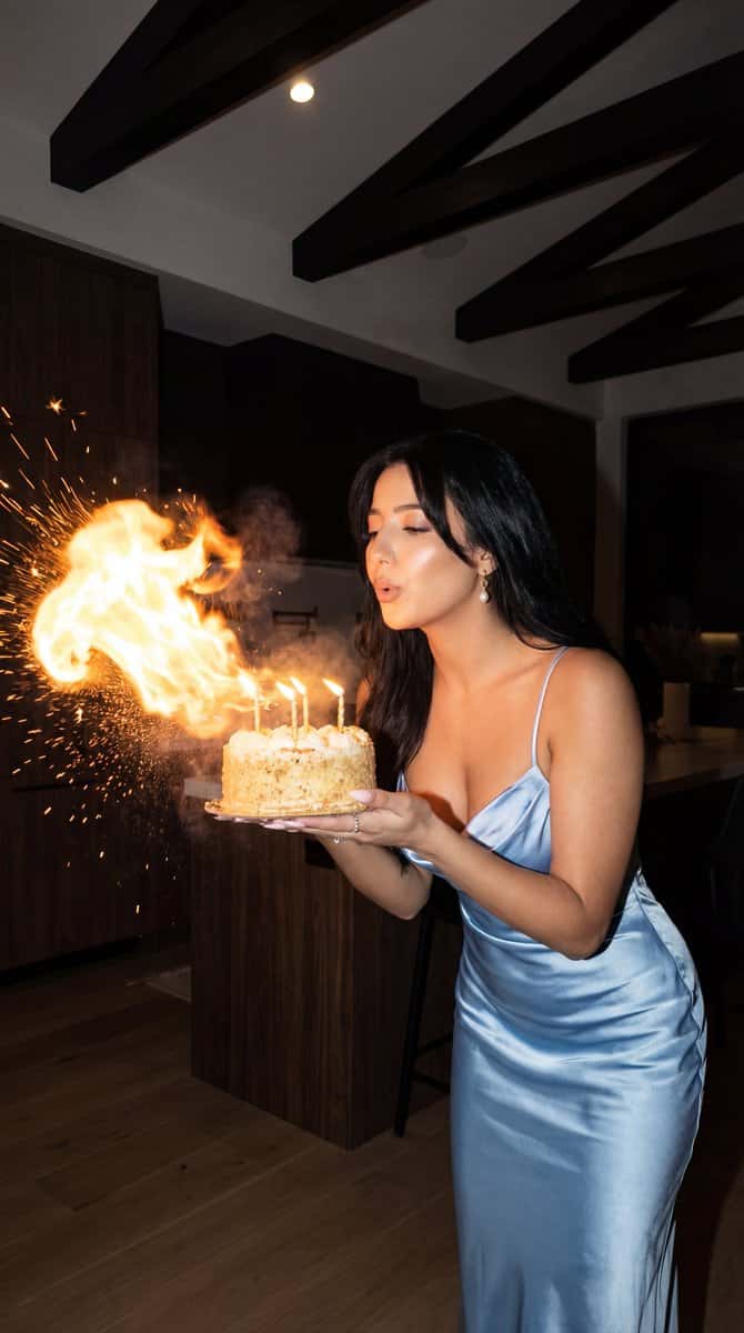 Ultra-photoreal candid birthday night portrait, 9:16 vertical. A woman in a shimmering light blue satin slip dress with thin straps stands in a dim modern interior with dark wood elements and geometric ceiling beams. She holds a small round birthday cake on a platter with both hands at chest height, leaning in to blow out the lit candles, lips pursed and cheeks slightly puffed. A dramatic flame burst erupts from the candles and expands to the left of the frame, with glowing sparks and glitter-like particles suspended in the air. Lighting: low ambient interior + warm candlelight highlights + subtle on-camera flash pop, realistic shadows, bright flame core, mild halation around highlights. Composition: subject on the right third, flame burst on the left third, background softly blurred. Photorealistic skin texture, no beautification, 35-50mm lens look, f/2.8, ISO 800, subtle film grain. No text, no watermarks.
