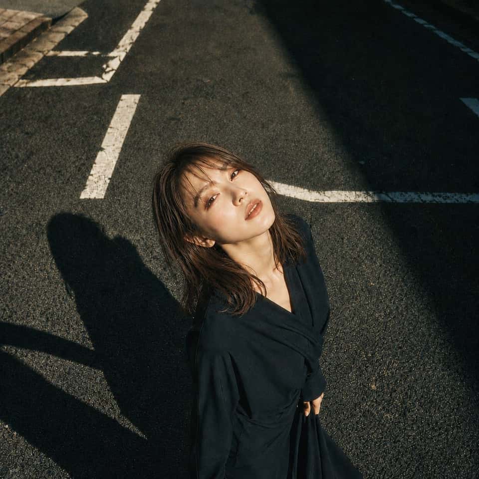 主題：白線と影 人物・表情：20代女性。明るいアイボリーベージュ寄りの白肌に、ほんのりピンクの血色を含んだ自然な肌質。頬、鼻先、鎖骨に柔らかなハイライト。精密で上品なドールライク日系メイク、淡いローズピンクのチーク、シャンパンベージュとピンクブラウンの目元、細く長いまつ毛、自然な涙袋、明るめブラウンの柔らかい眉、透け感のあるピンクベージュのグロスリップ。瞳に小さなキャッチライト。静かに見上げる、少し余韻のある表情。服装・ポーズ：深い黒に近いネイビーの和モダンなラップワンピース。長袖、重なりのあるVライン、腰で布を結んだようなドレープ、落ち感のある柔らかな生地。身体は画面下側に立ち、上半身を少し後ろへ預ける。顔はカメラへ向けて上げ、右手はスカートの布を軽く持ち、左腕は自然に体側へ落とす。姿勢は大人っぽく、静かな緊張感を残す。背景・光：日本の都市のアスファルト道路。白い道路標示が斜めに入り、画面左上に歩道の縁石が少し見える。強い午後の太陽が画面左上から差し、顔と髪、胸元を明るく照らす。人物の影は画面左下へ長く伸び、画面右上には建物の濃い影が大きく落ちる。白線、影、アスファルトの粒感をはっきり描写する。構図・カメラ：1:1の正方形。高い位置から見下ろす俯瞰ポートレート。人物は画面下中央寄りに配置し、道路の余白を広く残す。カメラは真上より少し斜め、人物の視線はレンズへ向かう。中距離のストリートポートレート、背景まで質感が残る自然な被写界深度。質感・スタイル：富士カメラのフィルムシミュレーションを思わせる写真感。クラシッククローム風の落ち着いた色調、やや抑えた彩度、暖かい肌色、少し冷たい深い影、柔らかなハイライトの粘り、繊細なフィルム粒子。過度なHDRや強いシャープネスは避け、リアルな日系ストリート写真として仕上げる。ネガティブ：不自然な顔や視線、幼い顔、過度な美肌補正、プラスチックのような肌、髪の塊感、余分な指、欠けた指、手足の融合、服の破綻、道路標示の文字化け、不要な人物や車、ロゴ、透かし、過度なHDR。