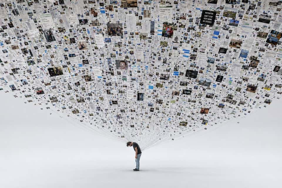 A person standing alone in an empty white void, but physically attached to them by thin threads of light are roughly 47,000 floating printed photographs — the entire contents of their phone's camera roll — most of them are screenshots of text messages, memes, receipts, photographs of parking spots, blurry nothing, duplicate duplicate duplicate, only a handful are actual meaningful moments (a dying grandmother smiling, a wedding kiss, a newborn) and these precious few are buried in the mass, the person is bent slightly under the weight, the image asks without asking: what are you actually documenting, hyperrealistic photography, studio lighting, conceptual art gravitas, Andreas Gursky scale.