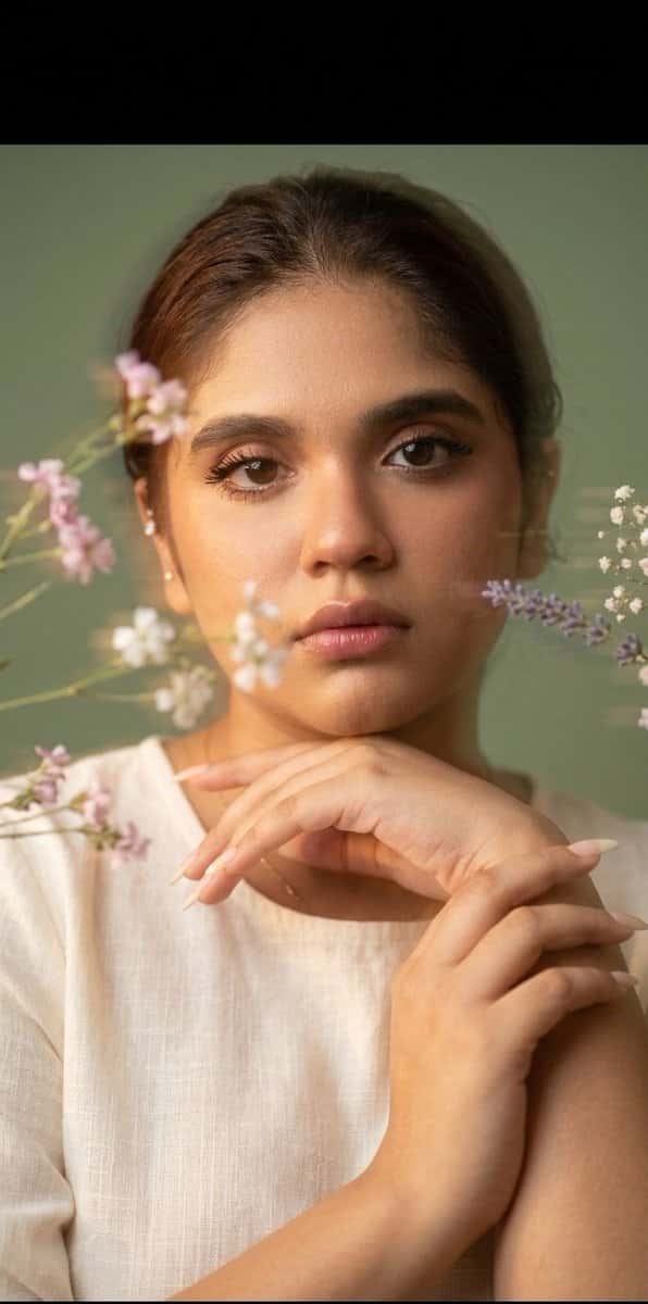 Ultra-realistic fine-art portrait of a young woman with soft natural features, minimal makeup, and calm introspective expression. Pastel muted background in sage green tones. Delicate flowers drifting across the frame in the foreground, creating intentional motion blur and soft streaks of light. Subtle double exposure and slow-shutter motion effects across the face, dreamy and poetic atmosphere. Gentle diffused lighting with smooth highlights on skin, natural texture preserved. Elegant minimalist composition, emotional and serene mood, editorial fine-art photography style, shallow depth of field, cinematic softness, painterly realism, ultra-high resolution, 8K quality.