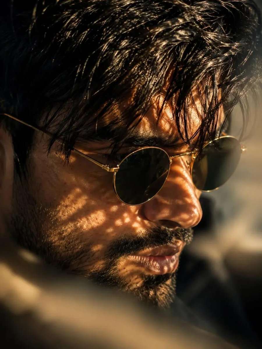 A sharp, low-angle side profile portrait of a handsome man with a thick, well-defined brown beard and mustache looking upward and toward the left. He has a messy dark hair and wearing thick-rimmed, polished black acetate sunglasses that sharp