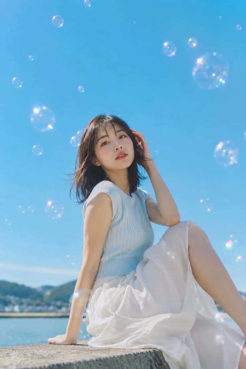 主題：夏泡の海辺 人物・表情：20代女性。自然で成熟した日系の顔立ち、清透感のある写真用メイク。肌は明るくなめらかだが、毛穴や産毛をわずかに残したリアルな質感。淡いローズ系チーク、細く自然なまつ毛、やわらかなブラウン眉、コーラルピンクのリップ。表情は落ち着いていて少し夢を見るように、唇を軽く開き、視線はカメラの少し上へ向ける。髪は暗めブラウンのミディアム、軽いシースルーバング、潮風で毛先と前髪が自然に揺れている。服装・ポーズ：淡いアイスブルーのノースリーブリブニット、白い軽やかなコットンロングスカート。清潔感のある夏の海辺コーデ。人物は海沿いの低いコンクリート壁に横向きで座り、上半身をカメラ側へ少しひねる。人物自身の右手は耳の横で髪を軽く押さえ、左手は壁につけて身体を支える。片膝を立て、もう片脚は画面右へ自然に流す。スカートは膝まわりで風を含み、布の重なりと接触影を出す。背景・光：湘南または鎌倉の海沿い。下部に海面、遠景にぼけた低い山並みと沿岸の街並み。画面の大部分は澄んだシアンブルーの空。大小のシャボン玉が人物の周囲と上空に浮かび、虹色の反射と柔らかなボケを作る。太陽光は画面左上から強めに入り、髪の輪郭、頬、肩、腕、スカート上部に明るいハイライト。影は画面右下へ浅く落ち、海からの反射光で肌をやさしく持ち上げる。構図・カメラ：縦長2:3。ローアングルの中景ポートレート。人物は画面下半分から中央に配置し、顔は中央より少し上。空の余白を大きく取り、シャボン玉で奥行きを作る。カメラは座面より低い位置から少し見上げる。広角すぎない自然な低角度で、脚や手が不自然に拡大しない。焦点は目元、背景の海と遠景は柔らかくぼかす。前景の泡は一部大きく淡くぼける。質感・スタイル：高品質な日系写真。夏の雑誌グラビアではなく、透明感のある日系ポートレート写真。原色を強くしすぎず、青空はシアン寄り、肌は明るく自然、白いスカートは飛ばしすぎない。光はまぶしいが硬すぎず、フィルム写真のような微細な粒子と淡いハレーションを加える。シャボン玉の虹色反射、布のしわ、髪の細い束、肌の自然な陰影を丁寧に描写する。ネガティブ：未成年に見える顔、幼態化、過度な美肌補正、プラスチックのような肌、安っぽい網紅風、過度な露出、透ける服、下着の露出、露点、不自然な顔や視線、余分または欠けた指、手足の融合、関節の破綻、脚の遠近感の崩れ、浮いた身体、服と身体の接触不良、光源と矛盾する影、泡の不自然な変形、文字、ロゴ、透かし、過度なHDR、過度なシャープ、明らかなAI感。