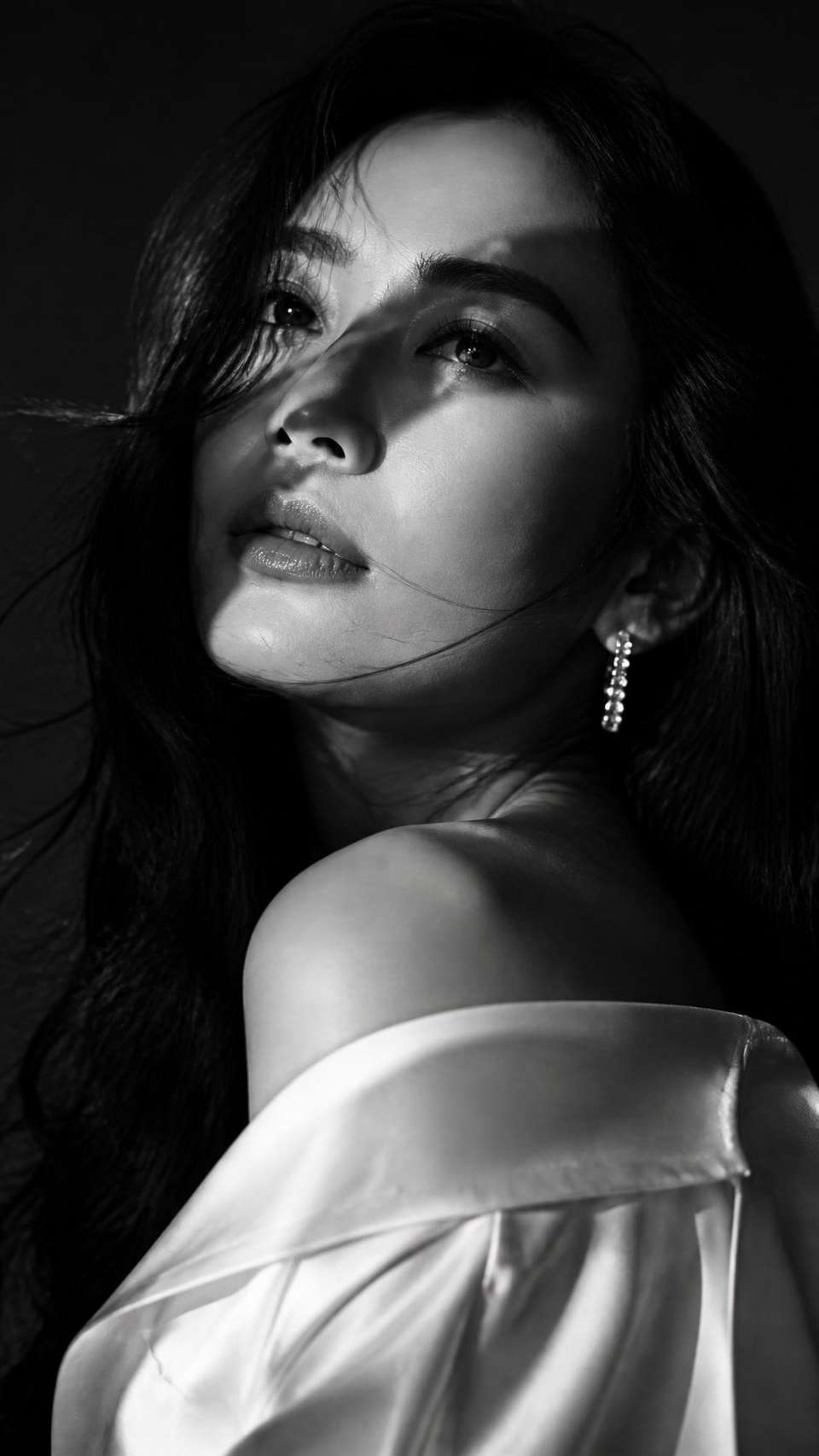 Ultra-realistic, cinematic, black-and-white Vogue editorial portrait of a beautiful Japanese woman. Three-quarter profile, shoulders turned away from camera, head gently tilted upward and looking off-frame with a calm, dreamy expression and softly parted lips. Long, naturally wavy black hair with loose strands flowing across her face. Elegant off-shoulder white satin blouse and delicate sparkling earrings. Dramatic hard side lighting casting bold diagonal shadows across eyes and face, high-contrast chiaroscuro, minimal dark studio background. 85mm lens, shallow depth of field, razor-sharp eyes, hyper-detailed natural skin texture, subtle film grain. Luxury fashion photography, timeless editorial aesthetic, ultra-photorealistic, 8K.