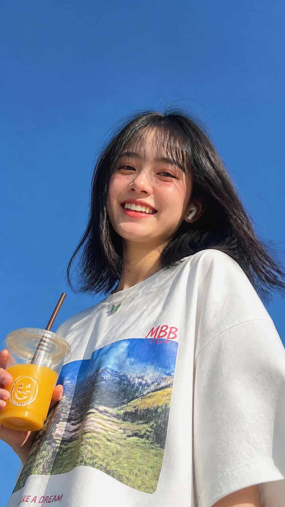 Ultra-realistic lifestyle portrait of a cheerful young woman with fair porcelain skin, medium-length straight black hair with wispy curtain bangs gently blown by the wind, bright genuine smile, almond-shaped dark brown eyes, and natural soft makeup. She wears an oversized white graphic T-shirt with colorful landscape prints, a delicate silver necklace with a small green butterfly pendant, and a white wireless earbud. She casually holds a transparent cup of iced orange juice with a brown straw and a cute smiling face logo. Dramatic low-angle wide-angle perspective looking up at her against a clear blue sky, illuminated by bright natural midday sunlight with soft shadows and vibrant colors. Fresh, youthful, carefree summer atmosphere, candid lifestyle photography, minimalist composition, natural skin texture, realistic hair strands, HDR, cinematic color grading, DSLR quality, sharp focus, ultra-photorealistic, editorial style, masterpiece, 8K.